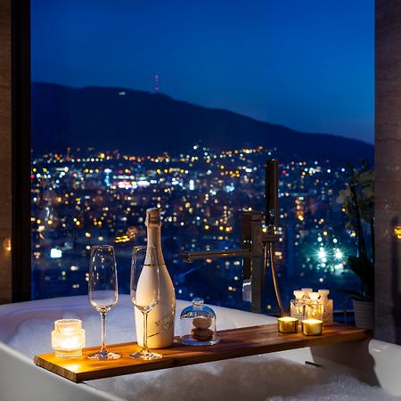 Grand Hotel Millennium Sofia Exterior photo The photo features a luxurious bathtub placed by a large window, showcasing a scenic city view at night. There is a wooden tray resting on the edge of the tub, displaying a bottle of champagne, two glass flutes, and several lit candles, creating an a