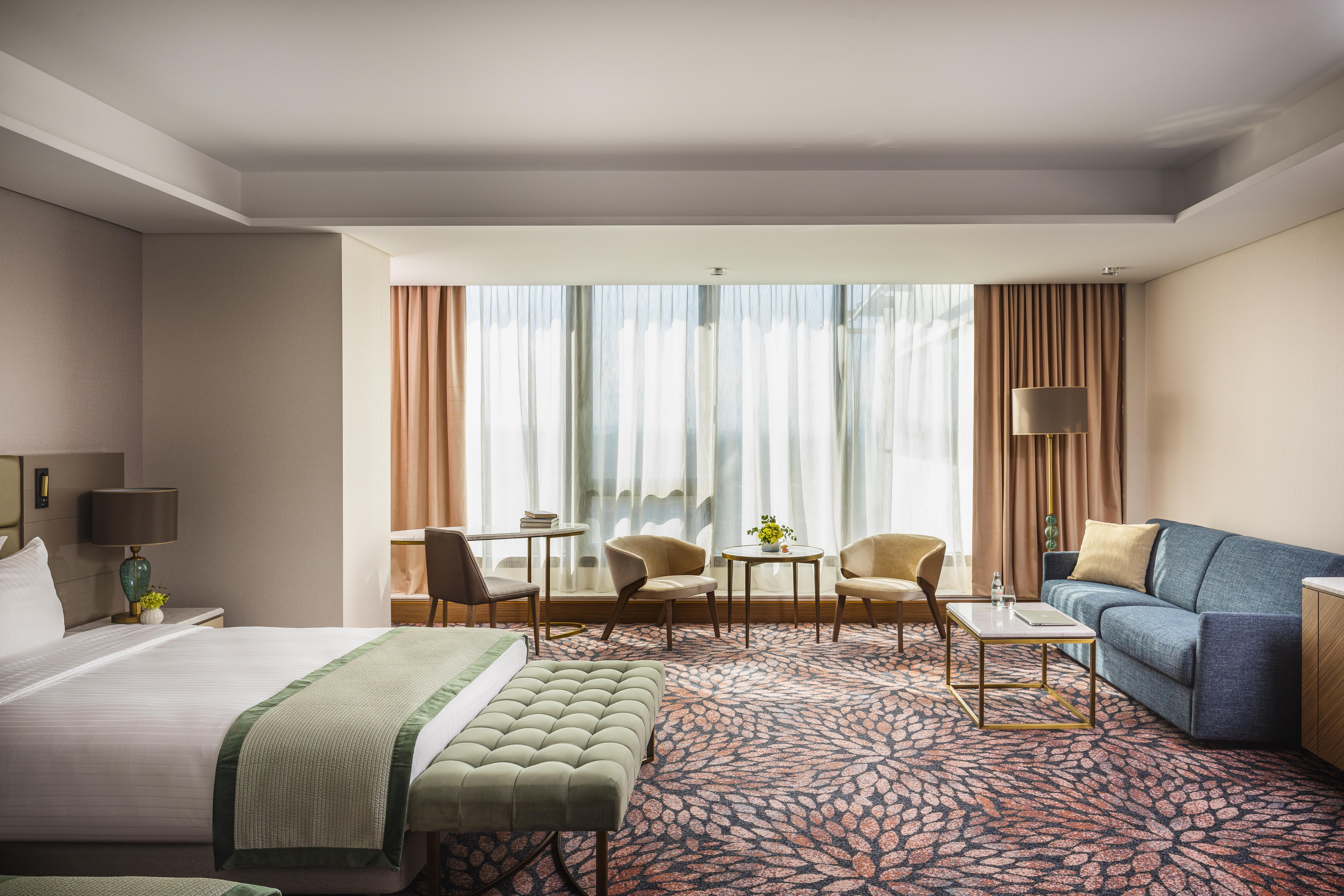 Grand Hotel Millennium Sofia Exterior photo The photo shows a modern hotel room featuring a spacious layout. On one side, there is a large bed with a green throw blanket and a tufted bench at its foot. The room is well-lit by large windows dressed with light curtains, which allow natural light