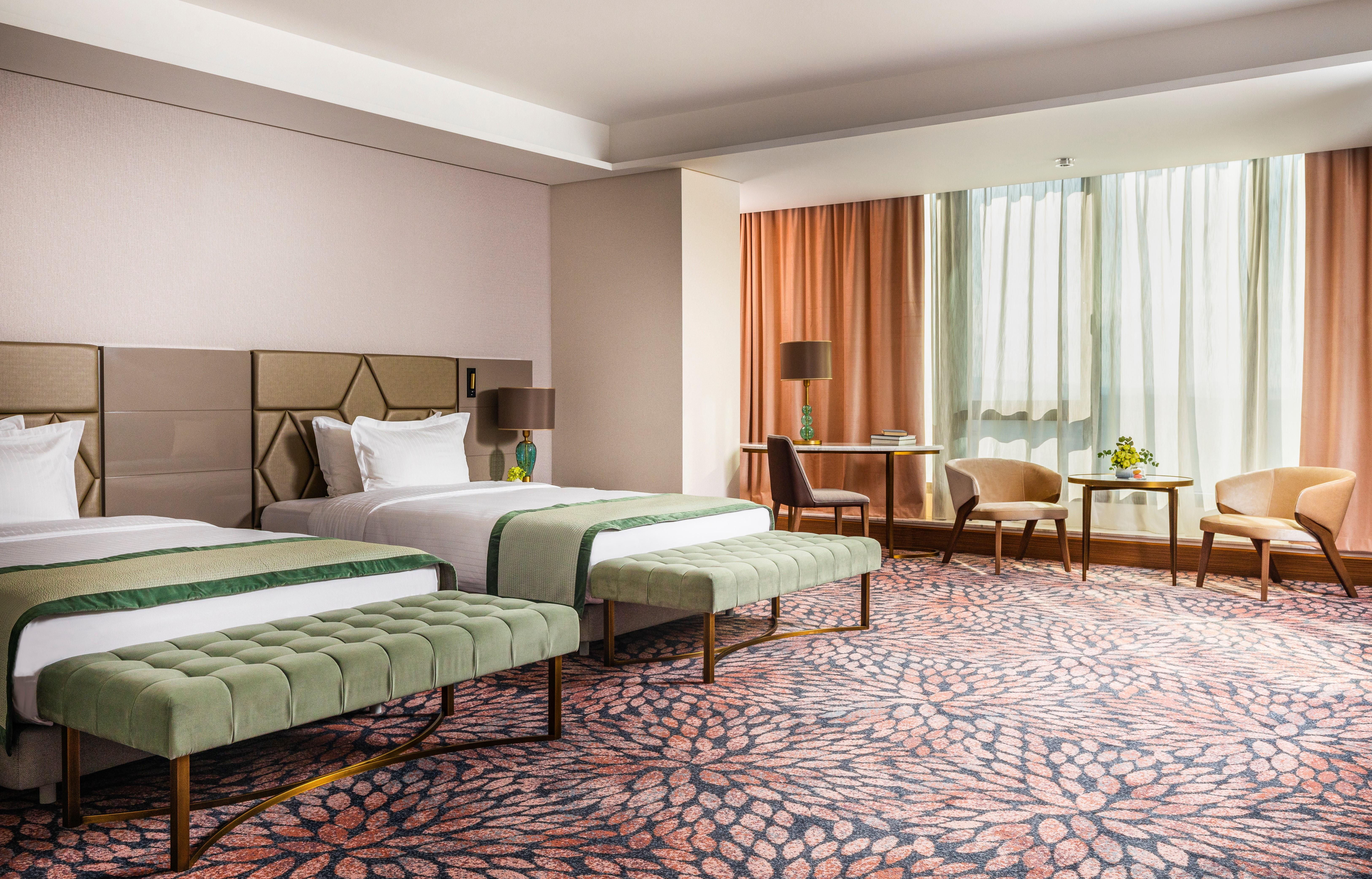 Grand Hotel Millennium Sofia Exterior photo The image shows a well-decorated hotel room featuring two beds with green striped bedspreads. There is a plush green bench at the foot of each bed. The room has large windows with sheer curtains allowing natural light to enter. There is a cozy seatin