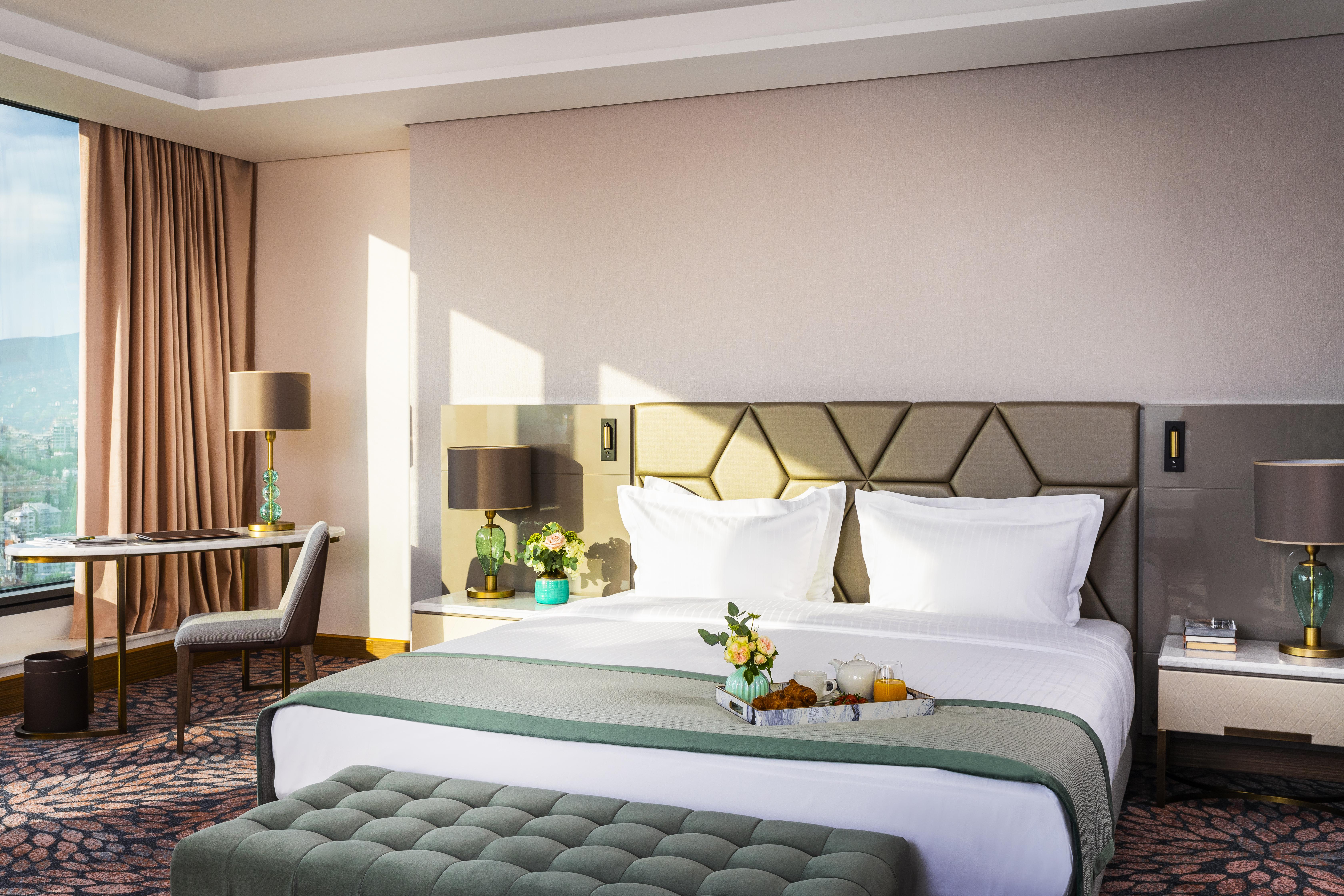 Grand Hotel Millennium Sofia Exterior photo The photo depicts a stylish hotel room featuring a large bed with white linens and a green decorative throw. Behind the bed is a geometric-patterned headboard. To the side, there are modern bedside lamps and a small table with a vase of flowers. A se