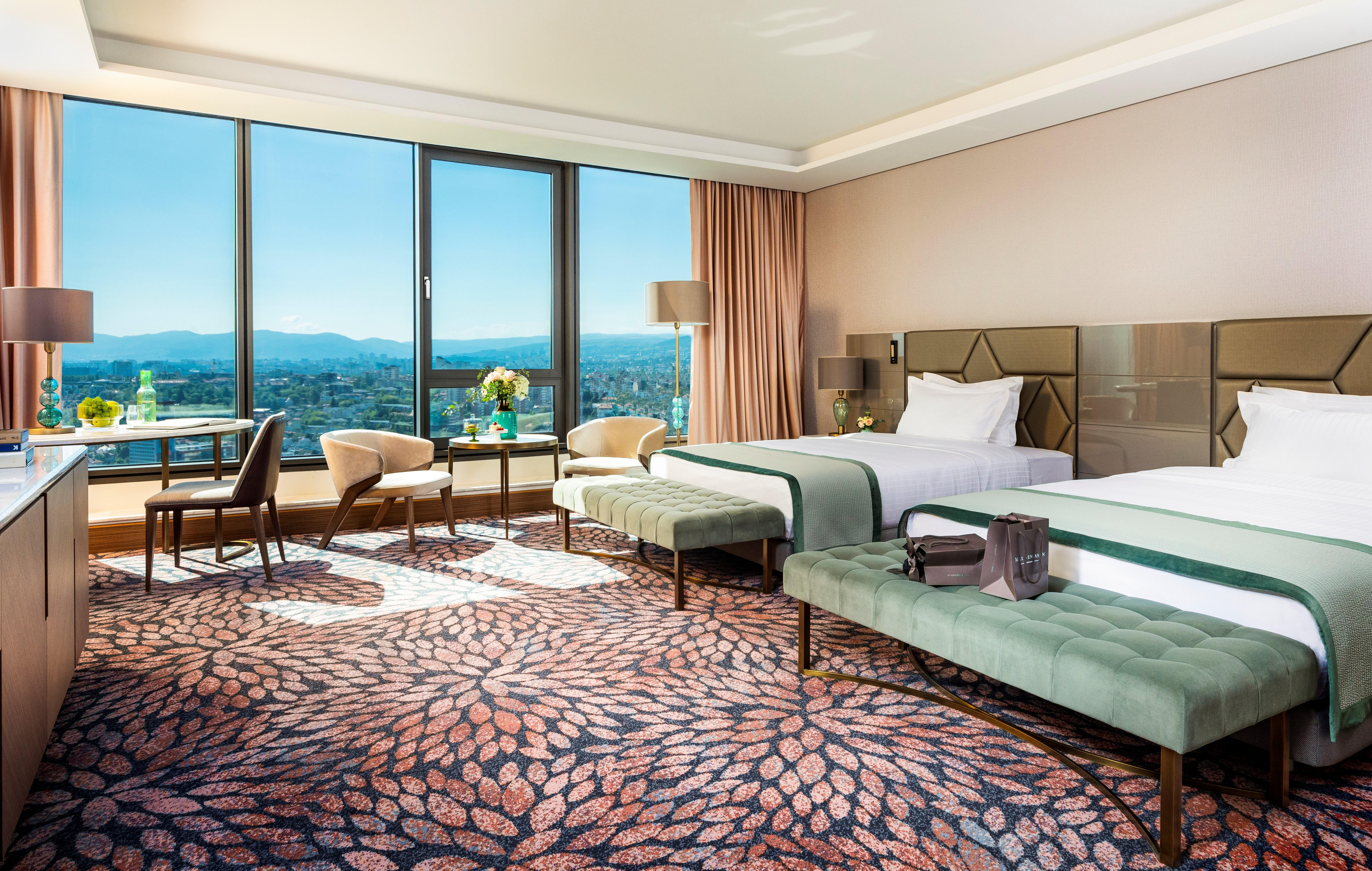 Grand Hotel Millennium Sofia Exterior photo The photo depicts a luxurious hotel room featuring two beds with elegant bedding. The room has large windows that offer a panoramic view of the city and mountains in the background. There is a decorative area rug with a floral pattern covering the fl