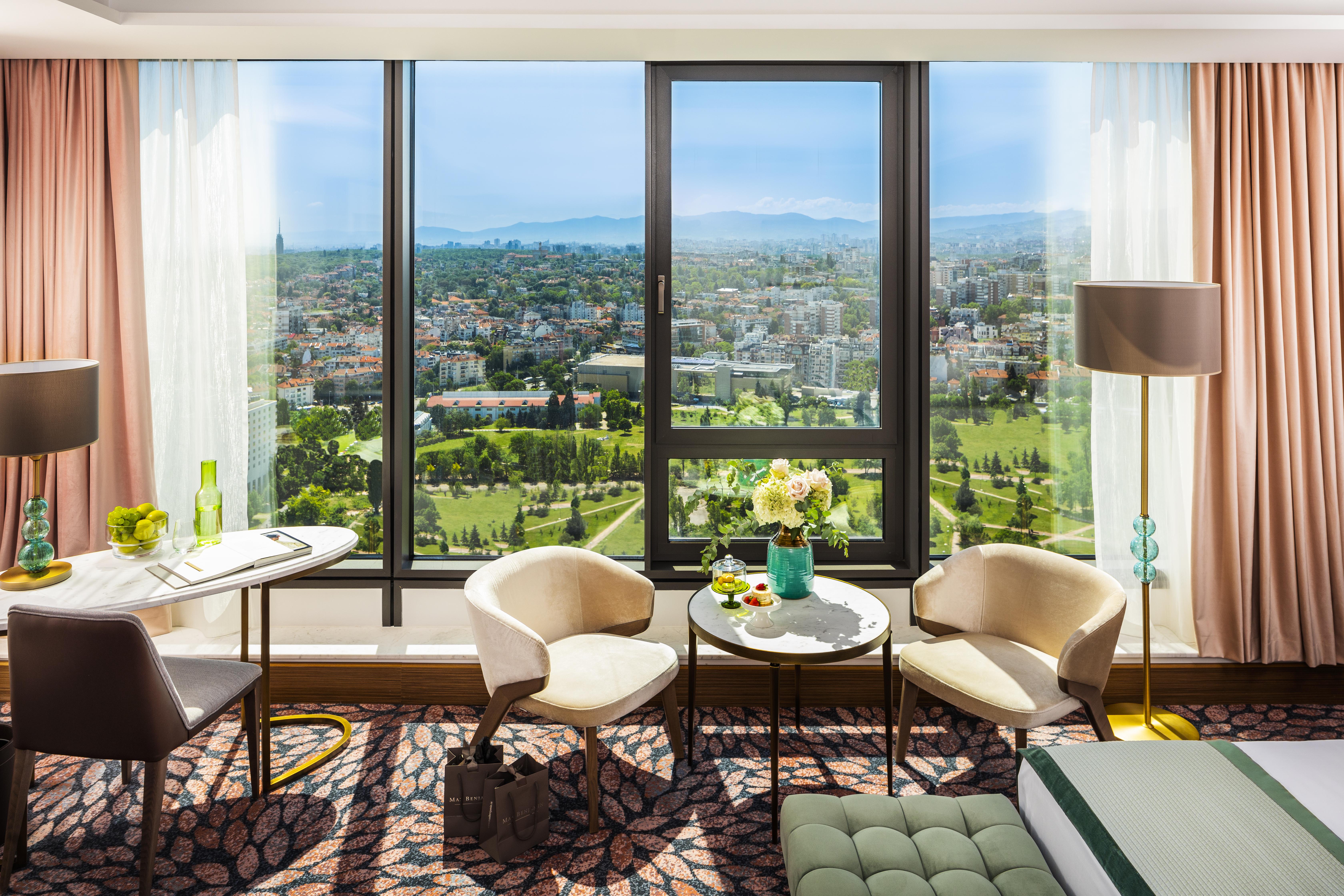 Grand Hotel Millennium Sofia Exterior photo The photo shows a stylish hotel room with large windows providing a beautiful view of a cityscape and landscape beyond. There are two modern chairs and a small round table set near the windows, accompanied by a vase of fresh flowers. The room feature