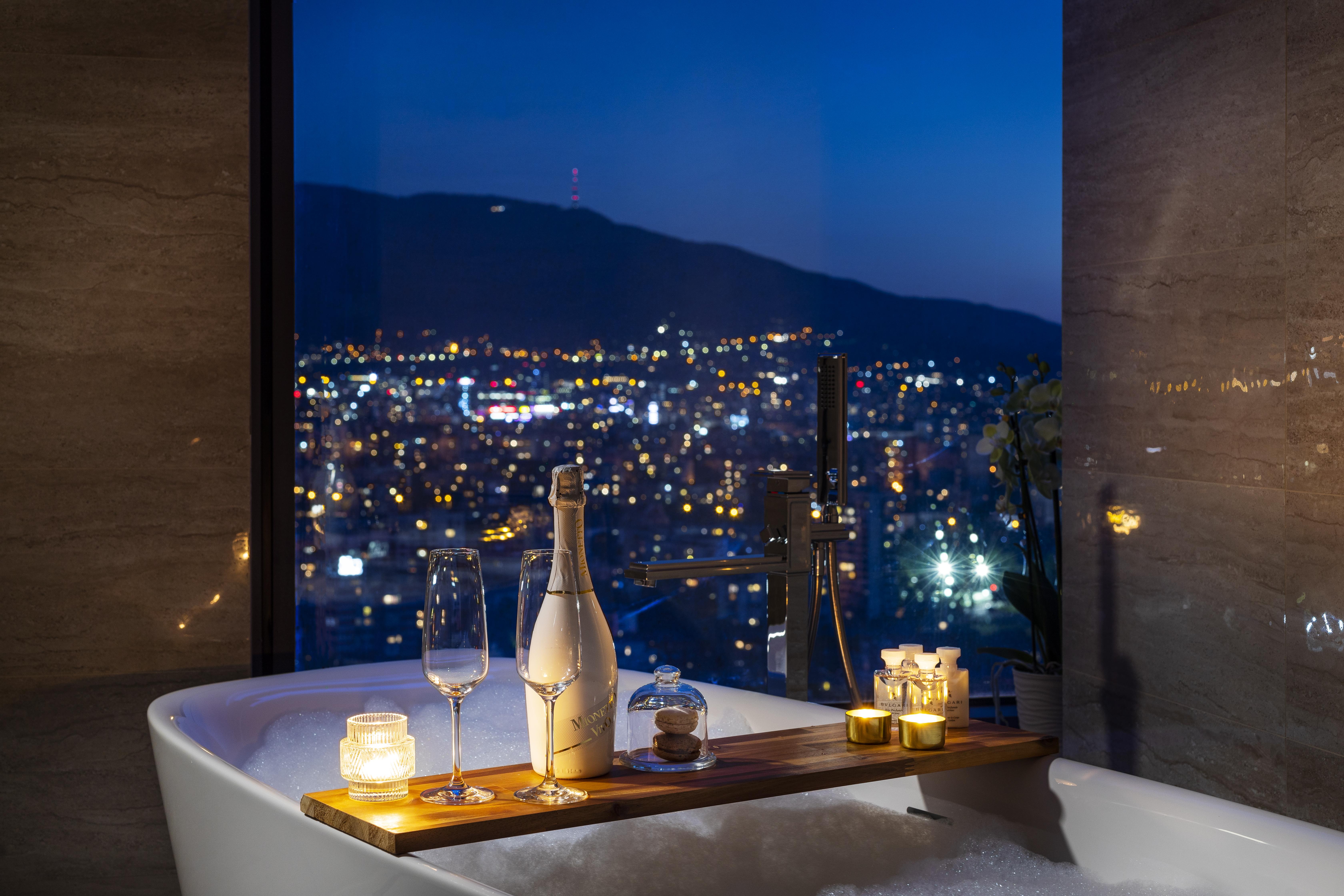 Grand Hotel Millennium Sofia Exterior photo The photo features a luxurious bathtub placed by a large window, showcasing a scenic city view at night. There is a wooden tray resting on the edge of the tub, displaying a bottle of champagne, two glass flutes, and several lit candles, creating an a