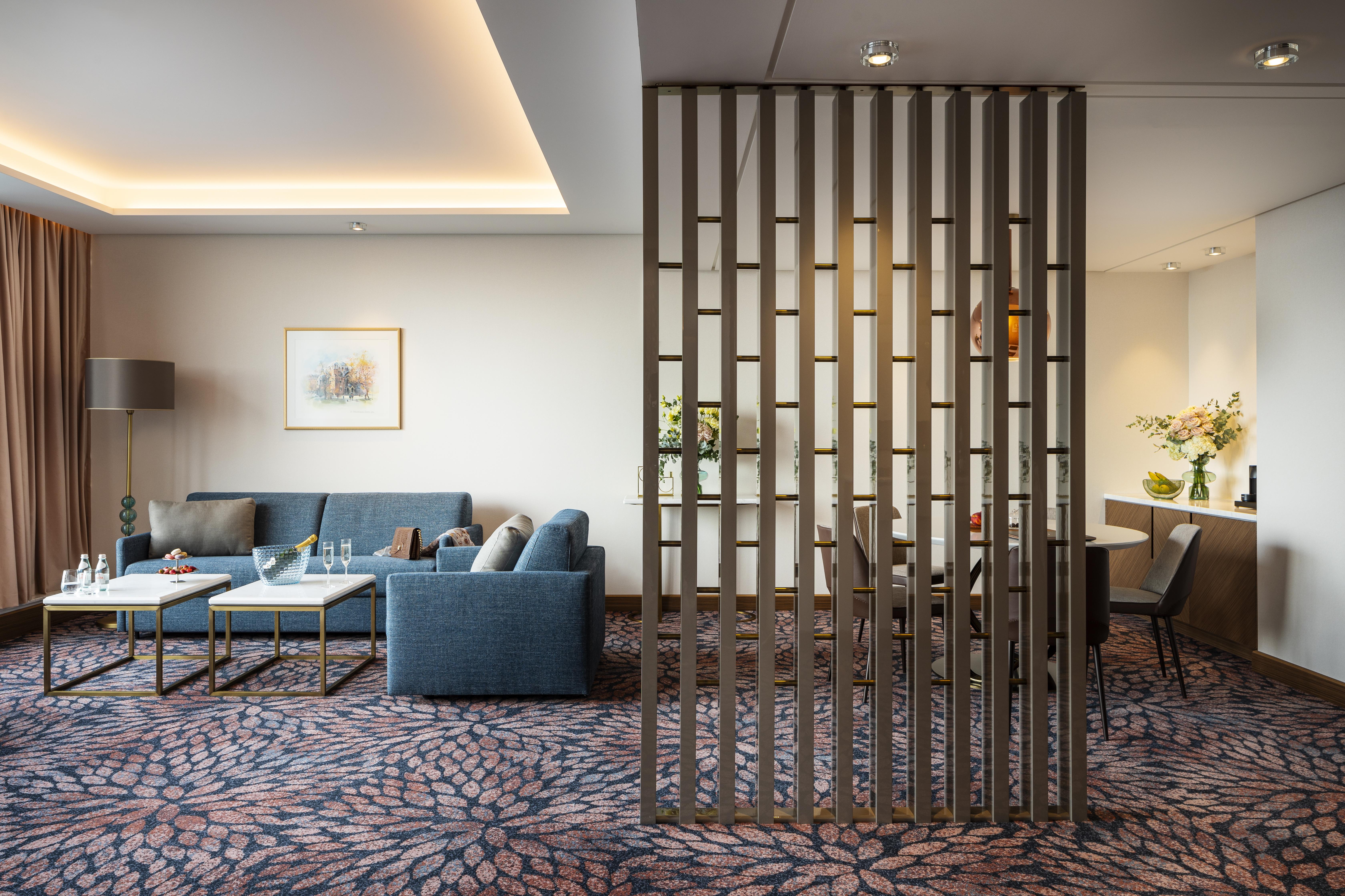 Grand Hotel Millennium Sofia Exterior photo The photo shows a modern living space with a comfortable seating area featuring a blue couch and two coffee tables. There is a decorative partition made of vertical wooden slats, which separates this sitting area from a dining space. The dining area 