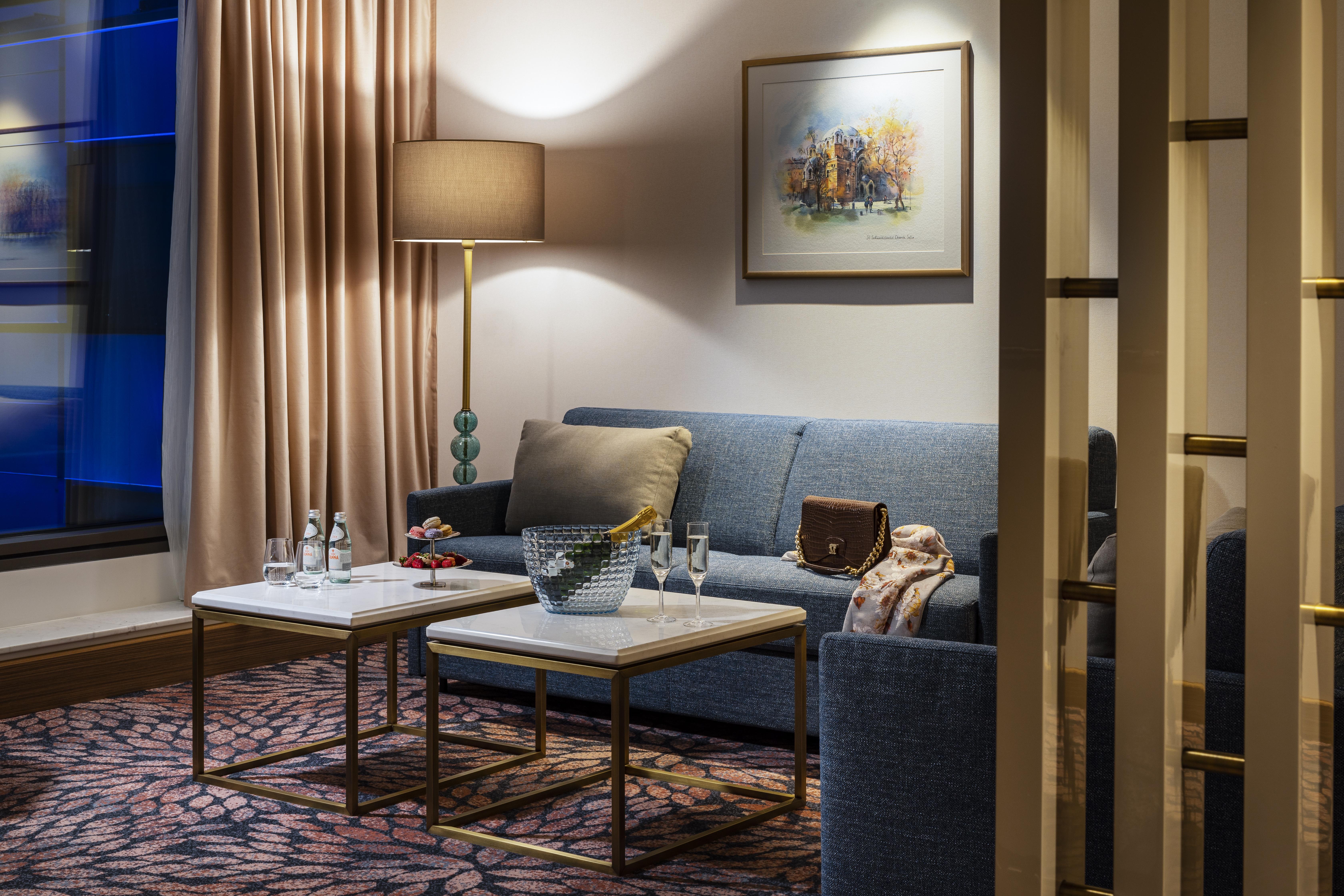 Grand Hotel Millennium Sofia Exterior photo The photo shows a cozy and elegantly decorated living space. It features a blue sofa against a wall, complemented by a painting above it. There are two small marble-top coffee tables with gold frames, one of which holds decorative items and glasses. 