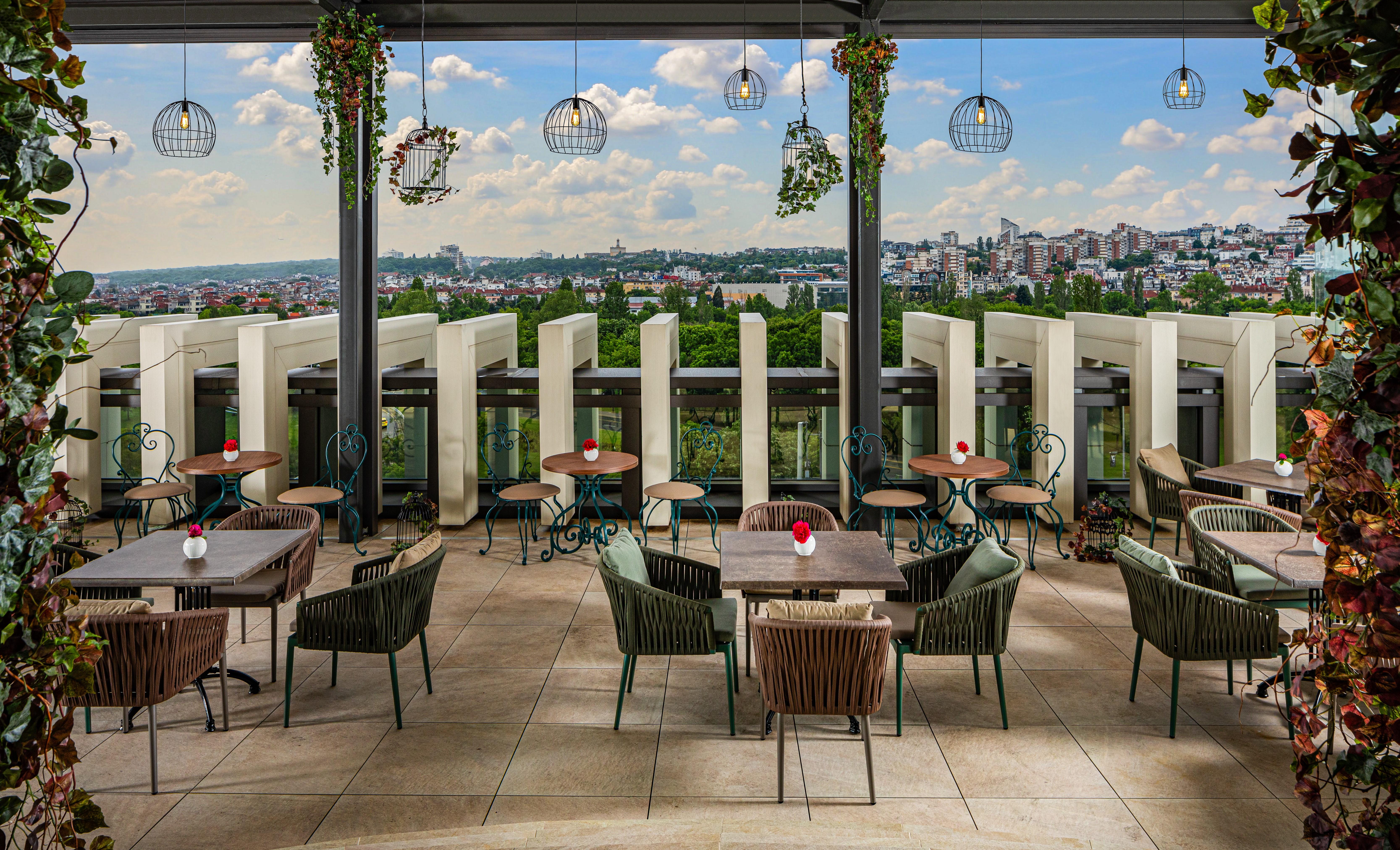 Grand Hotel Millennium Sofia Exterior photo The photo shows a stylish outdoor restaurant or café setting. There are several tables arranged on a tiled patio, each with chairs, some made of woven materials. Centerpieces with small red flowers adorn each table. The space is partially shaded by o