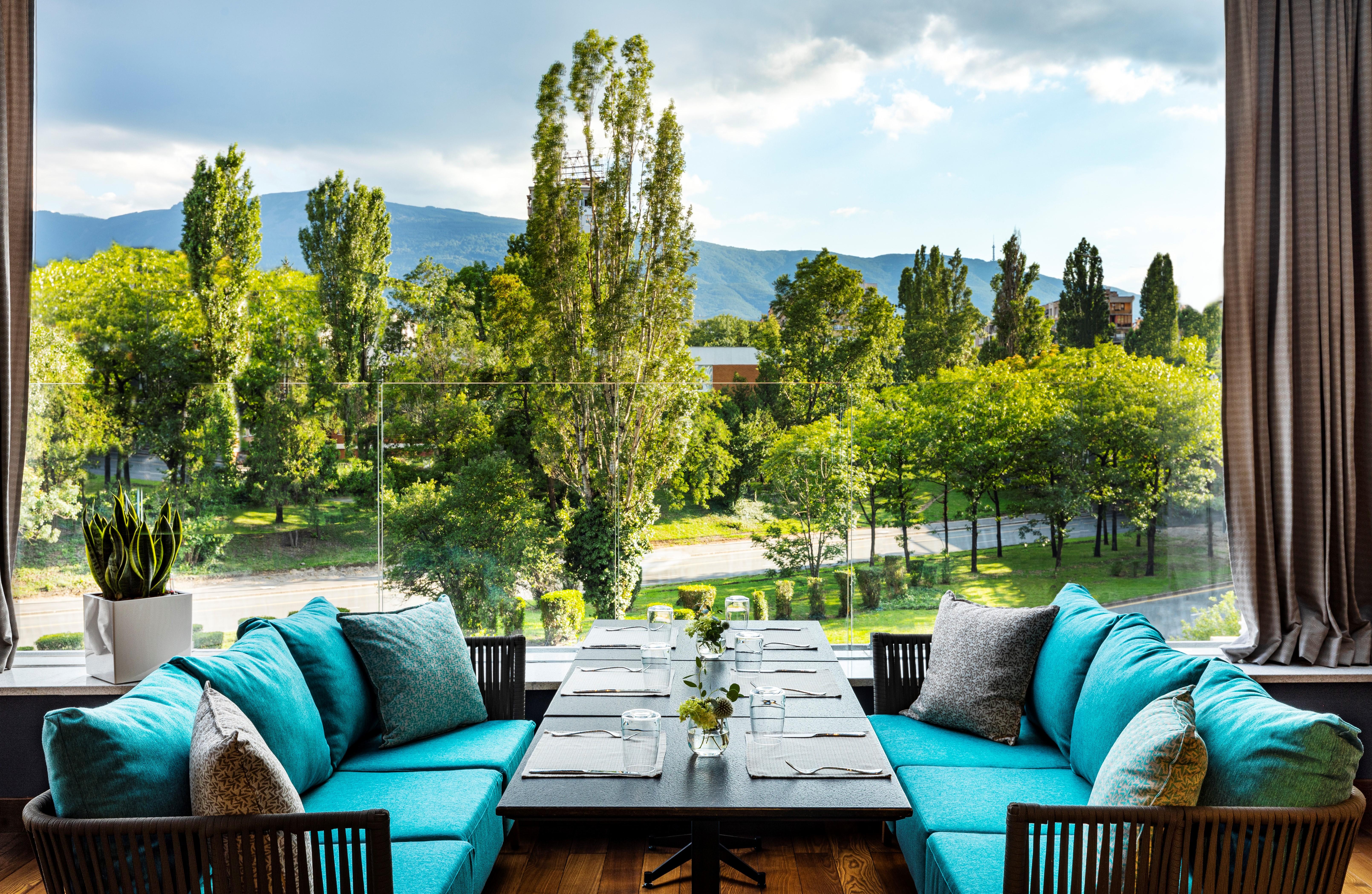Grand Hotel Millennium Sofia Exterior photo The photo shows a bright and inviting dining area with large windows that overlook a lush green landscape. There are several trees in the background, along with a view of distant mountains under a partly cloudy sky. The seating consists of stylish tu