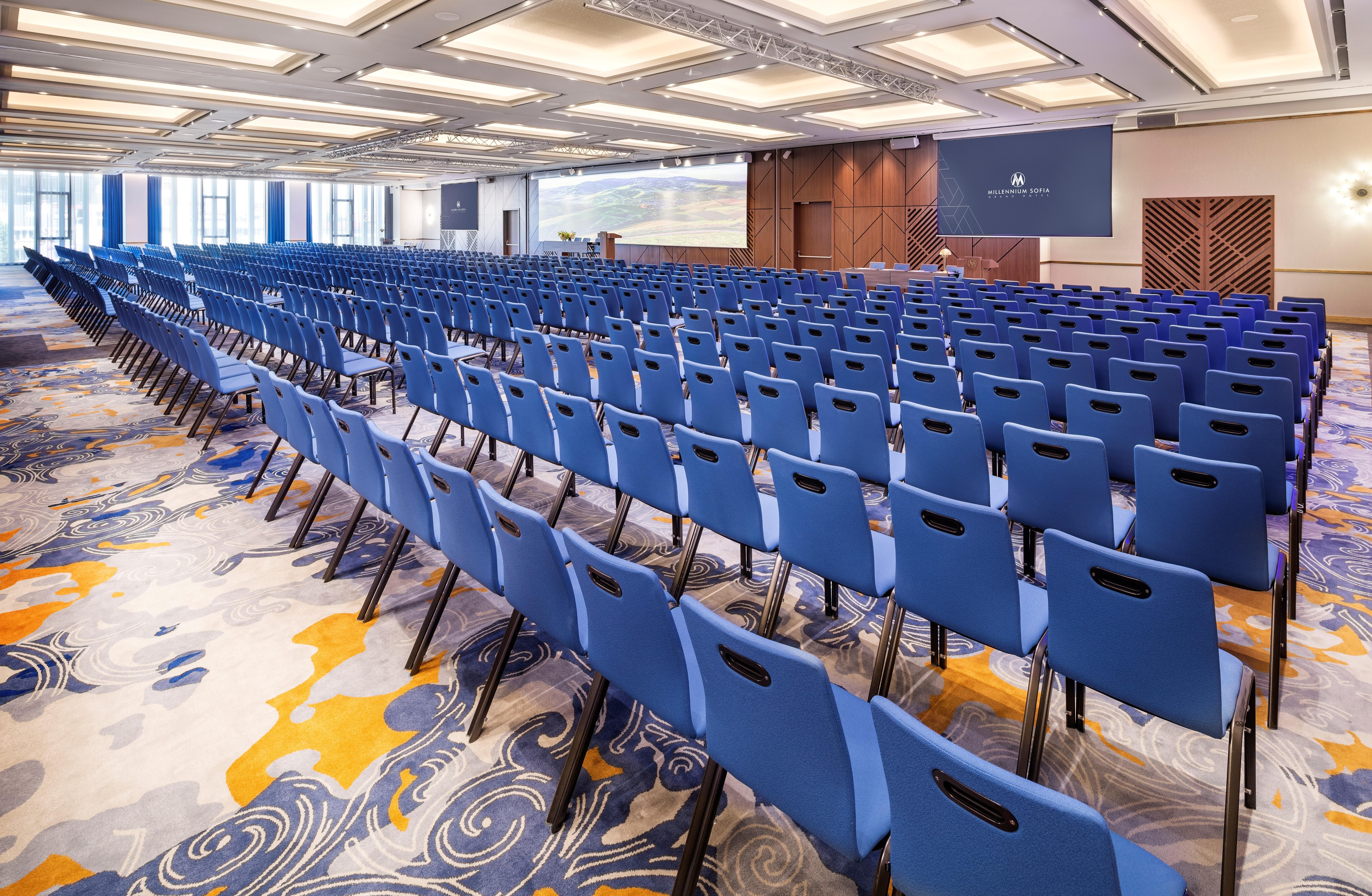 Grand Hotel Millennium Sofia Exterior photo The photo shows a large conference or meeting room set up for an event. There are numerous blue chairs arranged in rows, creating a seating area for attendees. The floor features a decorative pattern, and the room has a modern design with ample light