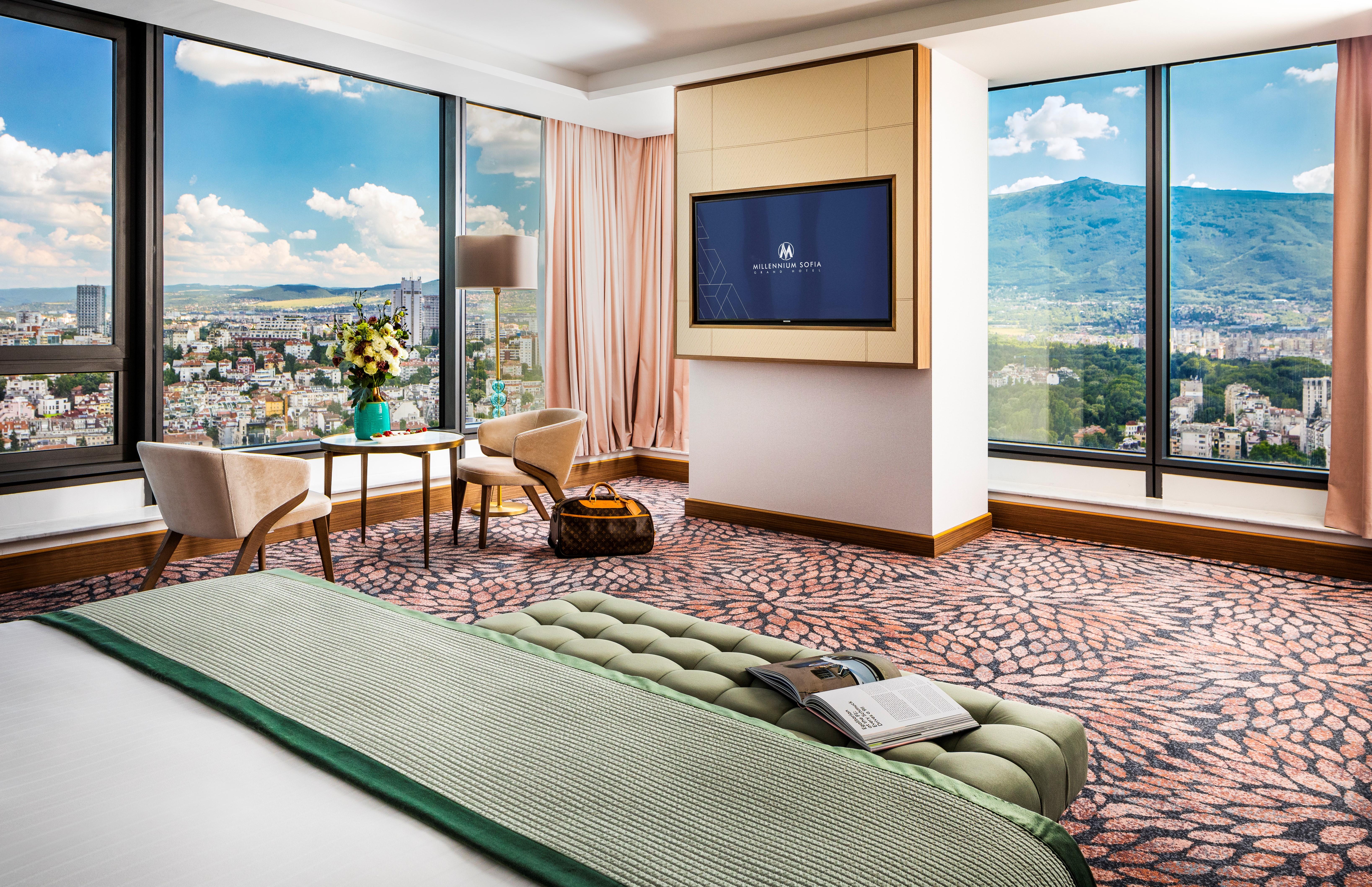 Grand Hotel Millennium Sofia Exterior photo The photo showcases a stylish hotel room with modern decor. It features large windows providing a panoramic view of a cityscape and mountains in the background. Inside, there is a comfortable bed with a green bedspread, a seating area with two armcha