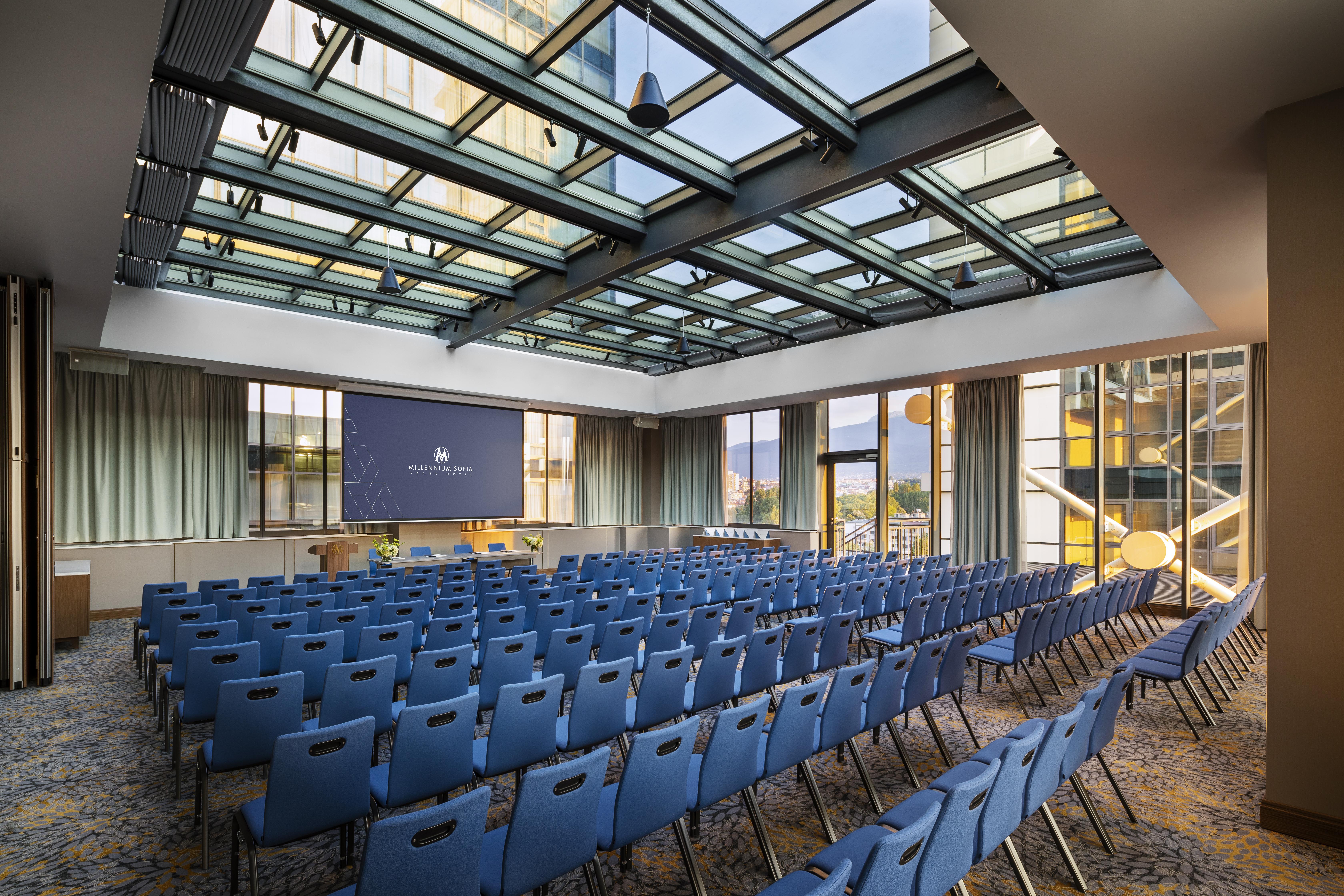 Grand Hotel Millennium Sofia Exterior photo The photo shows a large conference room or meeting space. It features rows of blue chairs arranged in a theater style, facing a large screen at the front of the room. The room is well-lit, with a glass ceiling allowing natural light to filter in. The