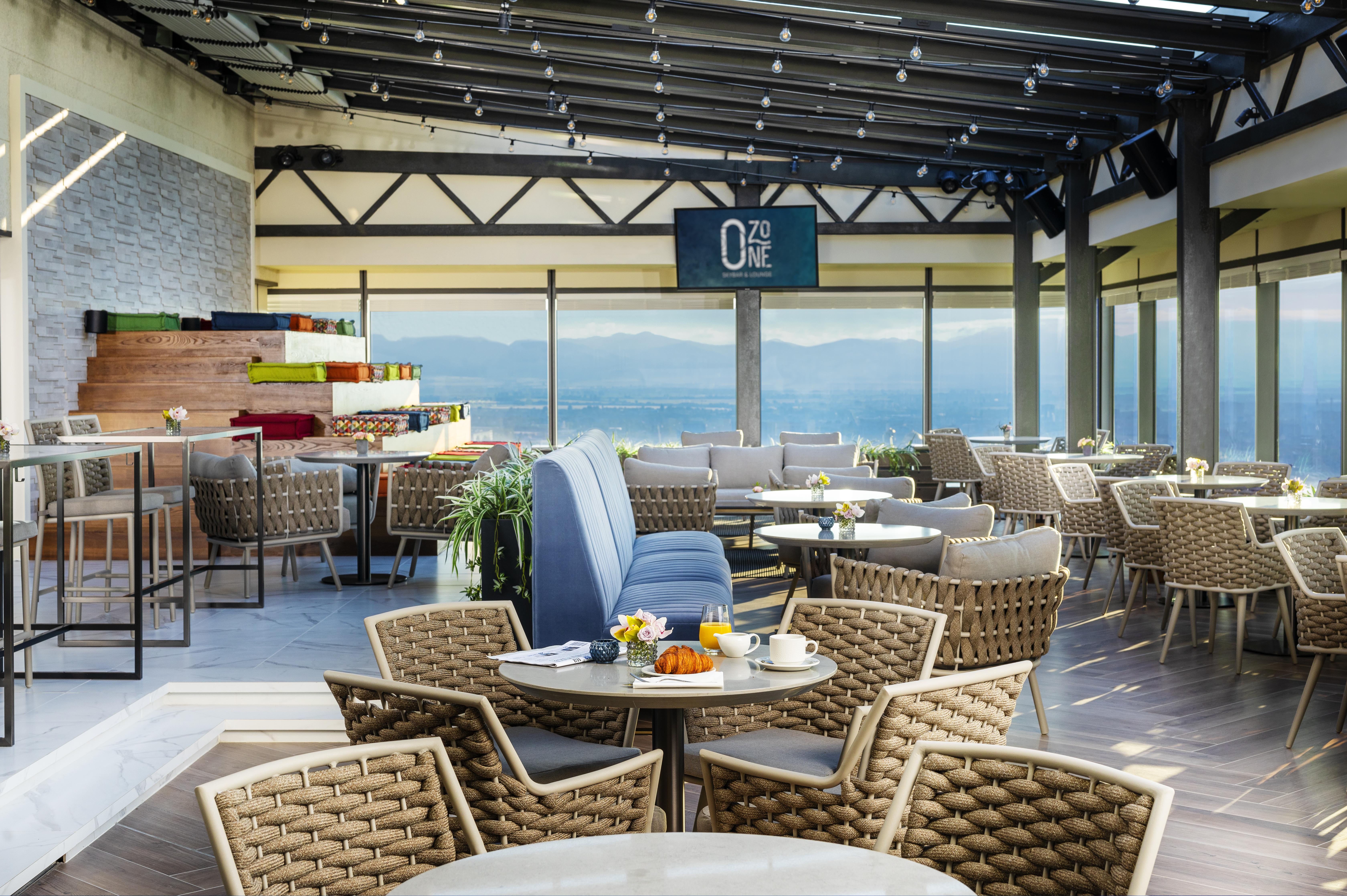 Grand Hotel Millennium Sofia Exterior photo The photo shows a modern restaurant or café with a spacious interior. It features a variety of seating arrangements, including round tables surrounded by wicker chairs and lounges. Large windows allow for a panoramic view, which likely includes sceni