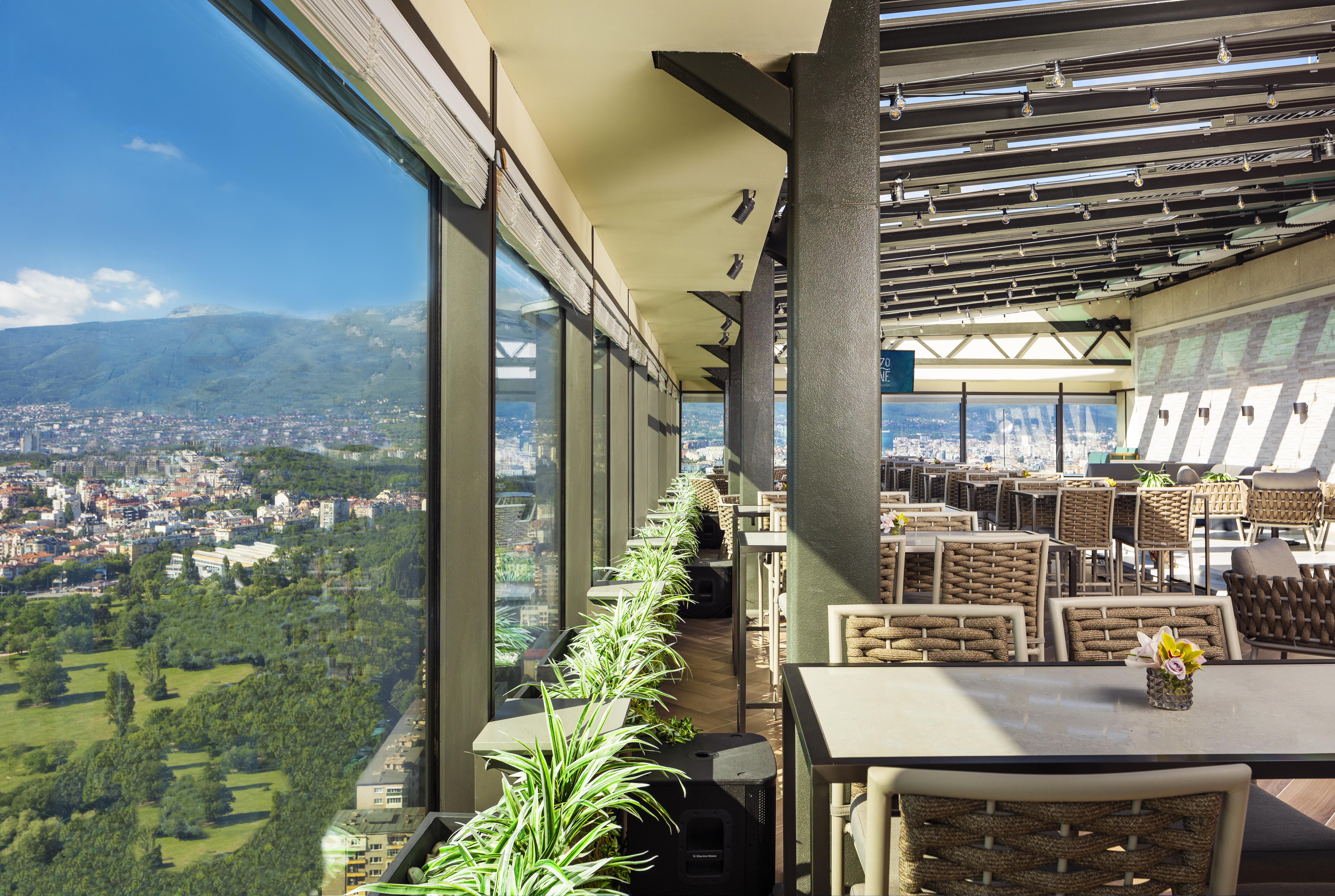 Grand Hotel Millennium Sofia Exterior photo The photo shows a modern, spacious restaurant with large glass windows offering a panoramic view of a city and surrounding landscape. The interior features contemporary furniture, including stylish tables and chairs. There are potted plants along the